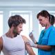 A female healthcare worker in blue scrubs and gloves administers an injection with a syringe to a man in a white tank top.