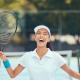 A young woman with a wide, joyful smile stands on an outdoor tennis court, holding a tennis racket in her right hand.