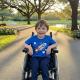 A young boy in a wheelchair smiles at the camera while sitting on a paved path in a park.