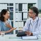 A woman in a light blue shirt talks to a male doctor in a white coat and stethoscope. The doctor is holding a tablet and listening to her. They are seated at a desk in an office.