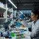 A female scientist in a lab coat and safety glasses examines a test tube in a laboratory.